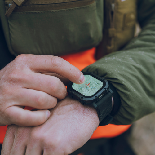 A close-up of a rugged black RECKON smartwatch with a compass-style face being touched by a finger, worn on a person's wrist. The person is wearing an olive green jacket and an orange safety vest, with a blurred outdoor background.