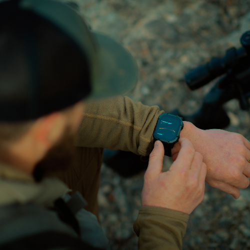 A close-up lifestyle shot of a man outdoors wearing a rugged black smartwatch with a dark teal screen, checking the time with his finger. He is wearing a tan long-sleeve shirt and a dark cap, with a blurred outdoor background.