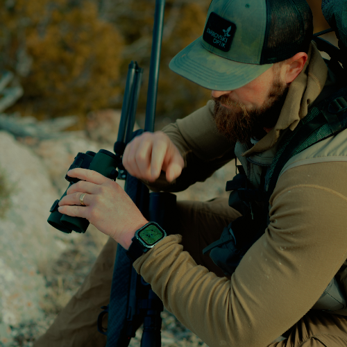 A bearded man in outdoor hunting attire, including a green cap with a 'Hunting Light' logo, a tan long-sleeve shirt, and a vest, is shown looking through binoculars while wearing a smartwatch.