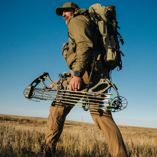 A man hiking through a grassy field carrying a compound bow and a large backpack under a clear blue sky.